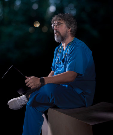 bearded mature doctor dressed in blue operating room suit sitting on a wooden box consulting a computer healthcare medical professionsの写真素材