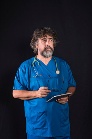 mature doctor with a beard dressed in a blue operating room suit on a black background consulting a...