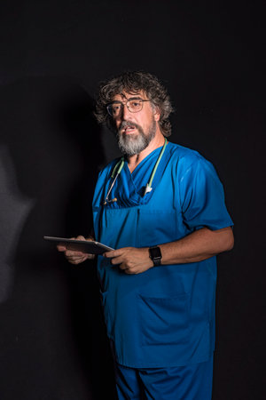 mature doctor with a beard dressed in a blue operating room suit on a black background consulting a tablet healthcare medical professionsの写真素材