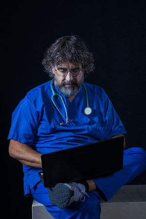 bearded mature doctor dressed in blue operating room suit sitting on a wooden box consulting a computer healthcare medical professionsの写真素材