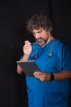 mature doctor with a beard dressed in a blue operating room suit on a black background consulting a tablet healthcare medical professionsの写真素材