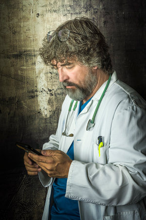 bearded mature doctor dressed in a blue surgical suit and a white coat looking at the mobile phone distractedly thinking about answering healthcare medical professionsの写真素材