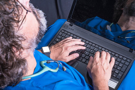 bearded mature doctor dressed in blue operating room suit sitting on a wooden box consulting a computer healthcare medical professionsの写真素材