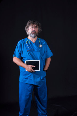 mature doctor with a beard dressed in a blue operating room suit on a black background holding a tablet looking at the camera healthcare medical professionsの写真素材