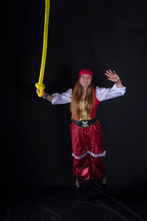 woman with long hair dressed as a pirate in a red suit and with a yellow balloon in the shape of a sword in her hand and on a black background photography of party eventsの写真素材