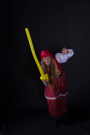 woman with long hair dressed as a pirate in a red suit and with a yellow balloon in the shape of a sword in her hand and on a black background photography of party eventsの写真素材