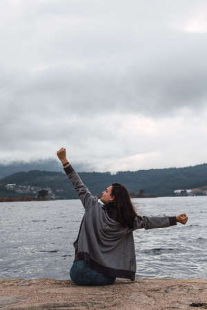 Woman is seeing and relaxing while she is sitting in fornt of the sea. She is rising her arms.の写真素材