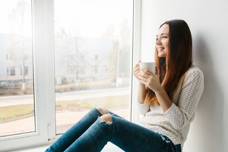 Beautiful young girl with a cup of tea sits by the windowの写真素材