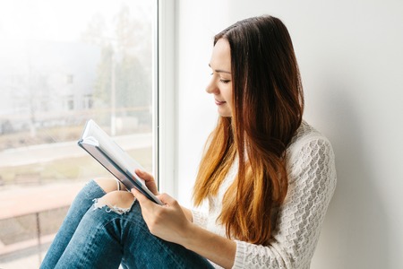 A pretty girl is reading a book sitting on the window. Obtaining knowledge, preparing for exams, students. Education and enjoyment of reading.の写真素材