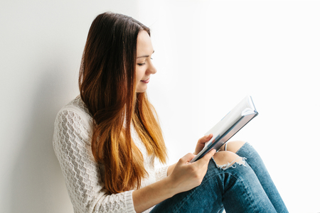 A pretty girl is reading a book sitting on the window. Obtaining knowledge, preparing for exams, students. Education and enjoyment of reading.の写真素材