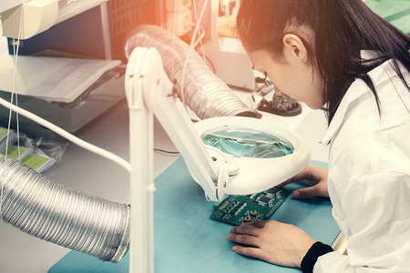 Beautiful female computer expert professional technician examining board computer in a laboratory in a factory. Troubleshooting. Technical support. Engineering. Manufacturing.の写真素材