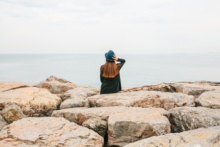 Young beautiful girl in a hat sits on the rocks on the seashore and looks into the distance. Rest, vacation, relaxation.の写真素材