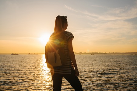 A young tourist girl with a backpack stands next to the sea at sunset and looks into the distance. Rest, relaxation, travel, vacation.の写真素材