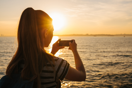 A young girl with a backpack pictures on the phone a beautiful view of the sea and the sunset. Picture for memory. Rest, travel, vacation.の写真素材