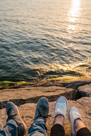 Male and female feet in sneakers on the rocky shore with beautiful view on wavy water, friendship or love concept. Trust and travel together.の写真素材