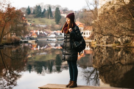 A tourist girl with a backpack standing on the shore and enjoying the nature and views of the city of Cesky Krumlov in the Czech Republic. The concept of solo travel.の写真素材