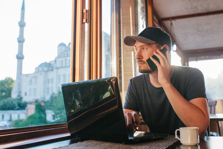 A young man-blogger freelancer working on a laptop in a cafe in Istanbul and calling by phone. View from the window to the world-famous Blue Mosque. Turkey.の写真素材