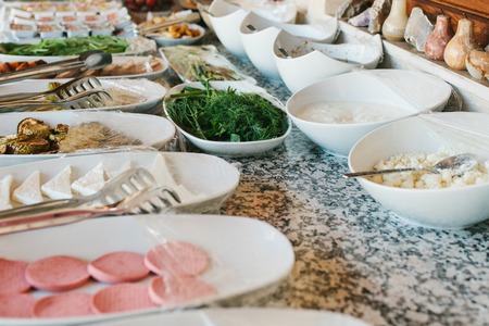Variety of fresh food on the table. Table with food: white plates and bowls with sausage sliced cheese and baked vegetables and herbsの写真素材