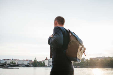 A tourist man with a backpack stands next to the river on the background of houses in Prague in the...