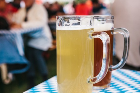 Two mugs with a light and dark beer stand on the table. In the background, blurred people. Celebrating the traditional German beer festival called Oktoberfestの写真素材
