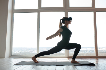 A young beautiful girl in virtual reality glasses makes yoga and aerobics remotely. Future technology concept. Modern imaging technology. Classes in single sports remotely.の写真素材
