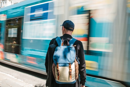 Man standing in front of road with tram. Male tourist in casual clothes with backpack is standing in front of road with tram is passing on sunny summer day. Move blur.の写真素材