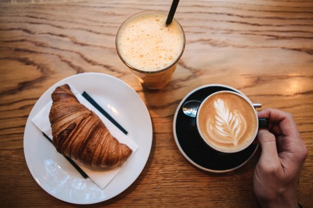 Top view. A man takes a cup of hot fragrant cappuccino. Near the table is a croissant and a glass with fresh orange juice.の写真素材
