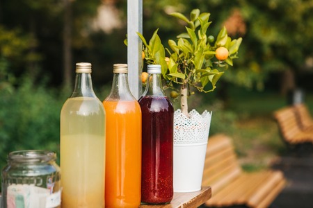 Selling refreshing homemade lemonade. Three bottles of lemonade stand in a row. Berry, orange and lemon with peppermint drinks inside the bottles. Benches in the park in the background.の写真素材