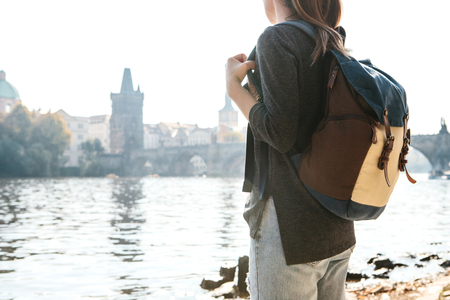 A beautiful young tourist girl with a backpack stands next to the Vltava river in Prague and admires one of the most popular attractions called the Charles Bridge. Travel around the Czech Republic.の写真素材