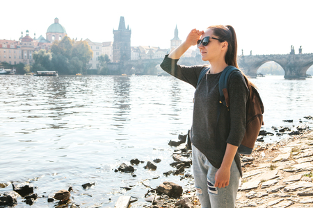 A beautiful young tourist girl with a backpack stands next to the river Vltava in Prague and admires the architecture of the city. Charles Bridge in the background. Travel around the Czech Republic.の写真素材