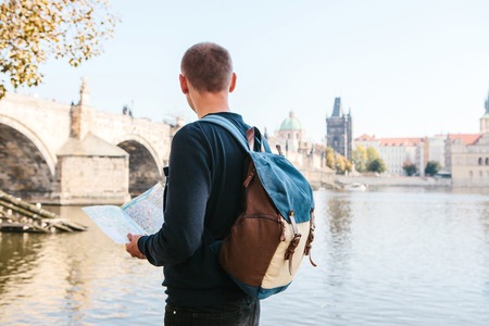 A young tourist man with a backpack standing next to the river Vltava in Prague looks at the map and admires the architecture of the city. Charles Bridge is near. Travel around the Czech Republic.の写真素材