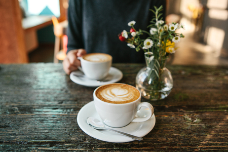 Two cups of coffee and a vase of flowers on a wooden table, the girl holds in her hand one cup of coffee in the background. A photo indicates a meeting of people and a joint pastime.の写真素材