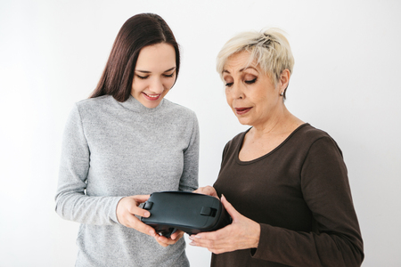 A young girl explains to an elderly woman how to use virtual reality glasses. The older generation and new technologies.の写真素材