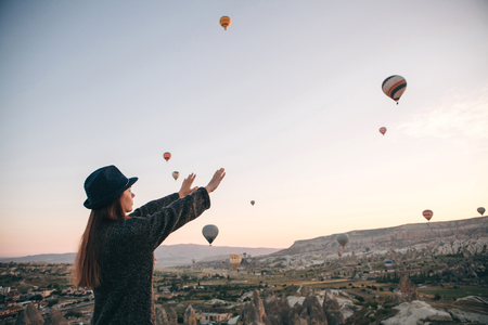 A tourist girl in a hat admires hot air balloons flying in the sky over Cappadocia in Turkey. Impressive sight.の写真素材