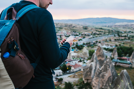 A tourist with a backpack on the background of the setting sun looks at the phone for geolocation and a mobile application and is going to call. In the background, the evening sunset and the city.の写真素材