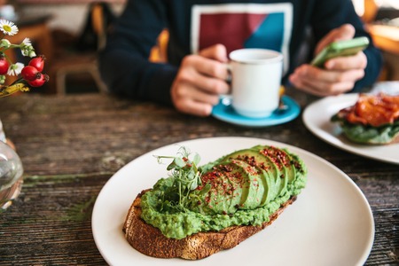 Sandwich with avacado on a wooden table. A man is going to call or write a message to another person in the background.の写真素材