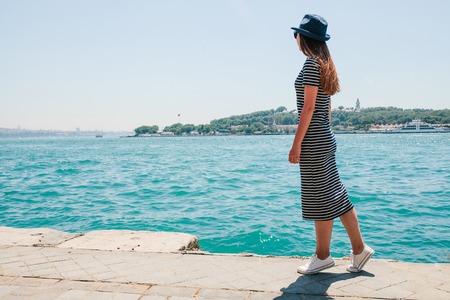 A young beautiful girl in a hat walks along the embankment and enjoys a holiday in Turkey and beautiful views of the Bosphorus and Istanbul in the distanceの写真素材