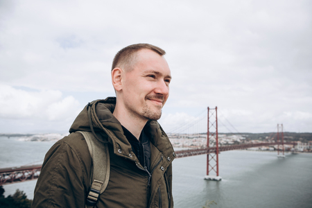 A young traveler or a tourist with a backpack on the waterfront in Lisbon in Portugal next to the 25th of April Bridgeの写真素材