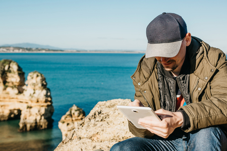 A young man or a tourist uses a tablet sitting on the Atlantic coast next to the city called Lagos in Portugal. Use of mobile applications or maps or freelancing or remote work or communication.の写真素材