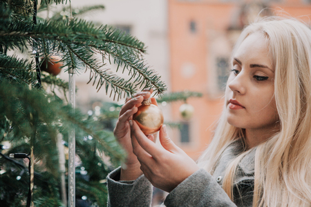 A young beautiful blonde woman touches a Christmas tree toy or ball or decorates a Christmas tree.の写真素材