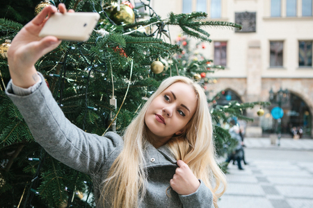 A beautiful young blonde woman or girl doing selfie or photographing next to a Christmas tree during Christmas holidays at the Old Town Square in Prague, Czech Republic.の写真素材