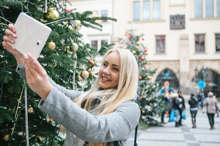 A beautiful young blonde woman or girl doing selfie or photographing next to a Christmas tree during Christmas holidays at the Old Town Square in Prague, Czech Republic.の写真素材