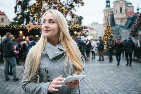 A beautiful young blonde woman or tourist uses a tablet next to a Christmas tree during the Christmas holidays at the Old Town Square in Prague, Czech Republic.の写真素材