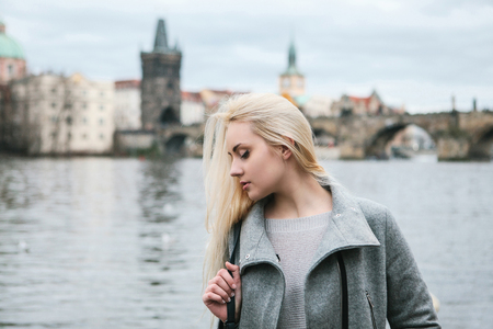 Fashionable blonde with long hair in a coat near Charles Bridge in Prague. Beautiful young woman outdoors.の写真素材