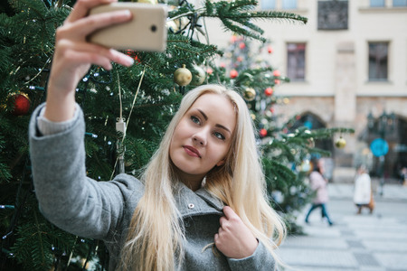 A beautiful young blonde woman or girl doing selfie or photographing next to a Christmas tree during Christmas holidays at the Old Town Square in Prague, Czech Republic.の写真素材