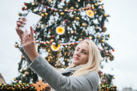 A beautiful young blonde woman or girl doing selfie or photographing next to a Christmas tree during Christmas holidays at the Old Town Square in Prague, Czech Republic.の写真素材