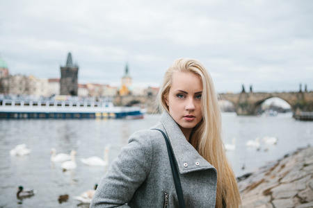 Fashionable blonde with long hair in a coat near Charles Bridge in Prague. Beautiful young woman outdoors.の写真素材