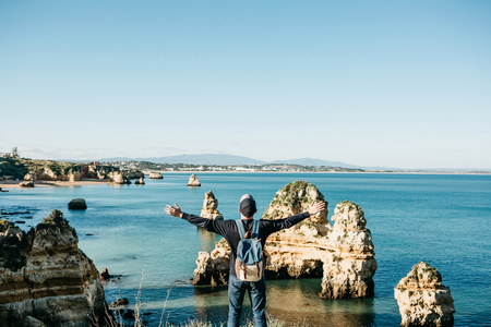 A tourist or traveler with a backpack admires the beautiful view of the Atlantic Ocean and the coast near the city called Lagos in Portugal and raises his hands up showing how he is free and he likes.の写真素材