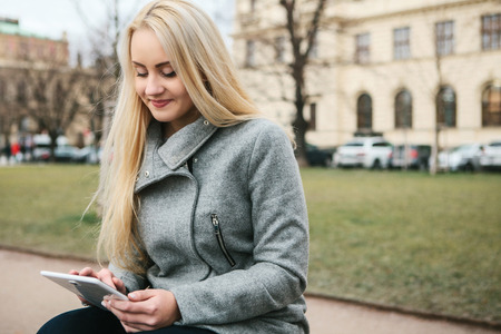 A beautiful blonde girl in a autumn coat sits on a street in Prague and uses a tablet to communicate with friends or for the Internet or reading a book or another.の写真素材