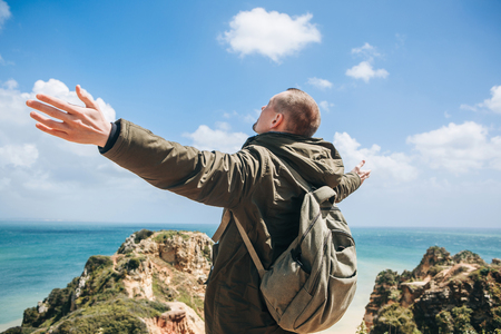 View from the back. A young man tourist enjoys the beautiful views of the Atlantic Ocean and the landscape off the coast in Portugal and raises his hands upward showing how pleased he is.の写真素材